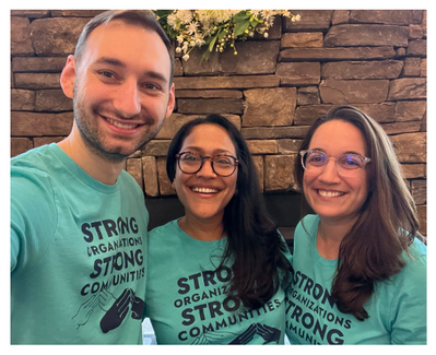 DOF team members Dan, Sasha, and Avi have their arms around each other’s shoulders and are smiling. They’re wearing “Strong Organizations, Strong Communities” teal t-shirts.