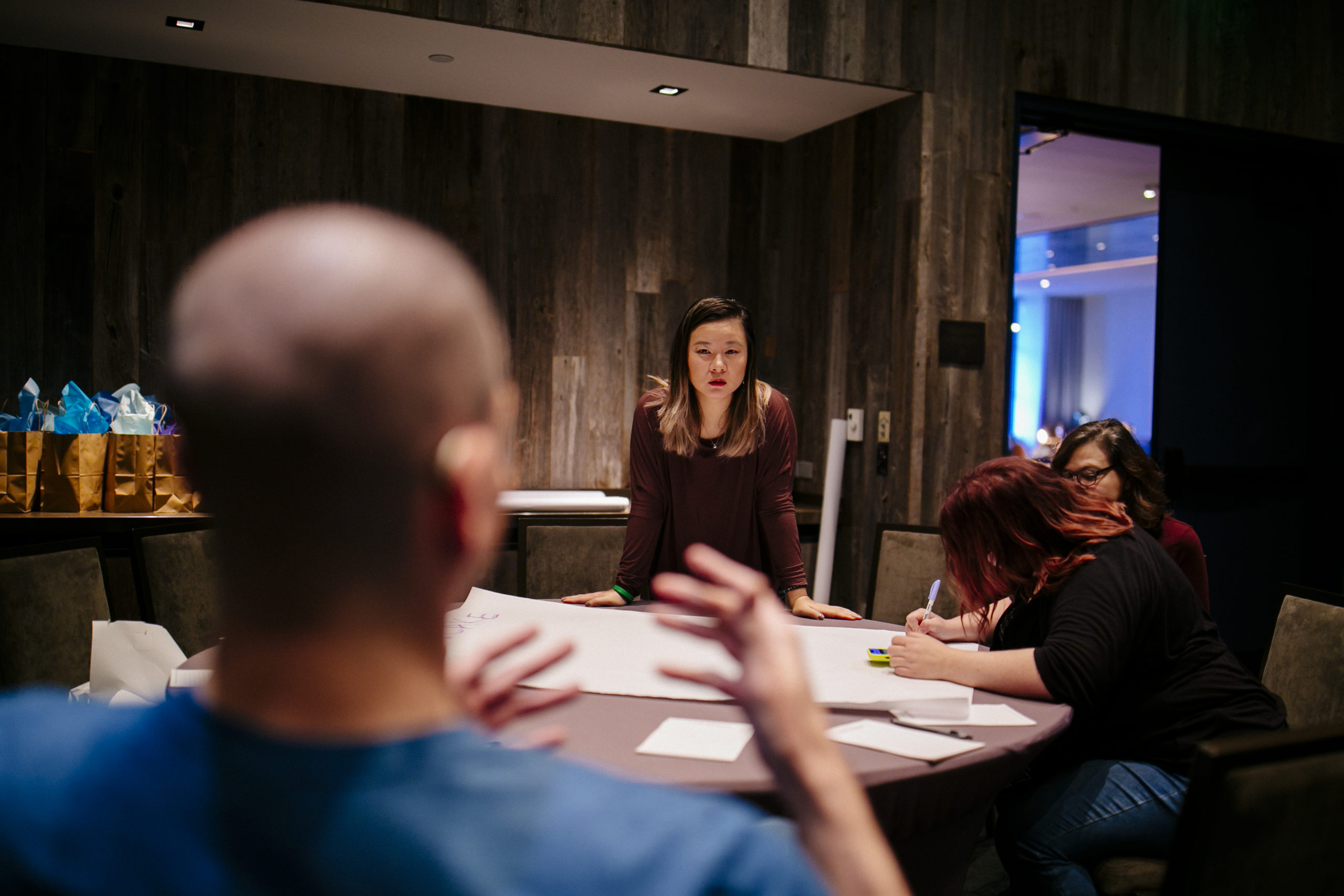 A group of four people sit around a table in a dimly lit room, engaged in discussion. A woman in a maroon shirt leans forward on the table, looking attentive, while another woman writes on a large sheet of paper. A man in the foreground gestures with his hands, and another woman sits nearby listening. Gift bags are visible in the background.
