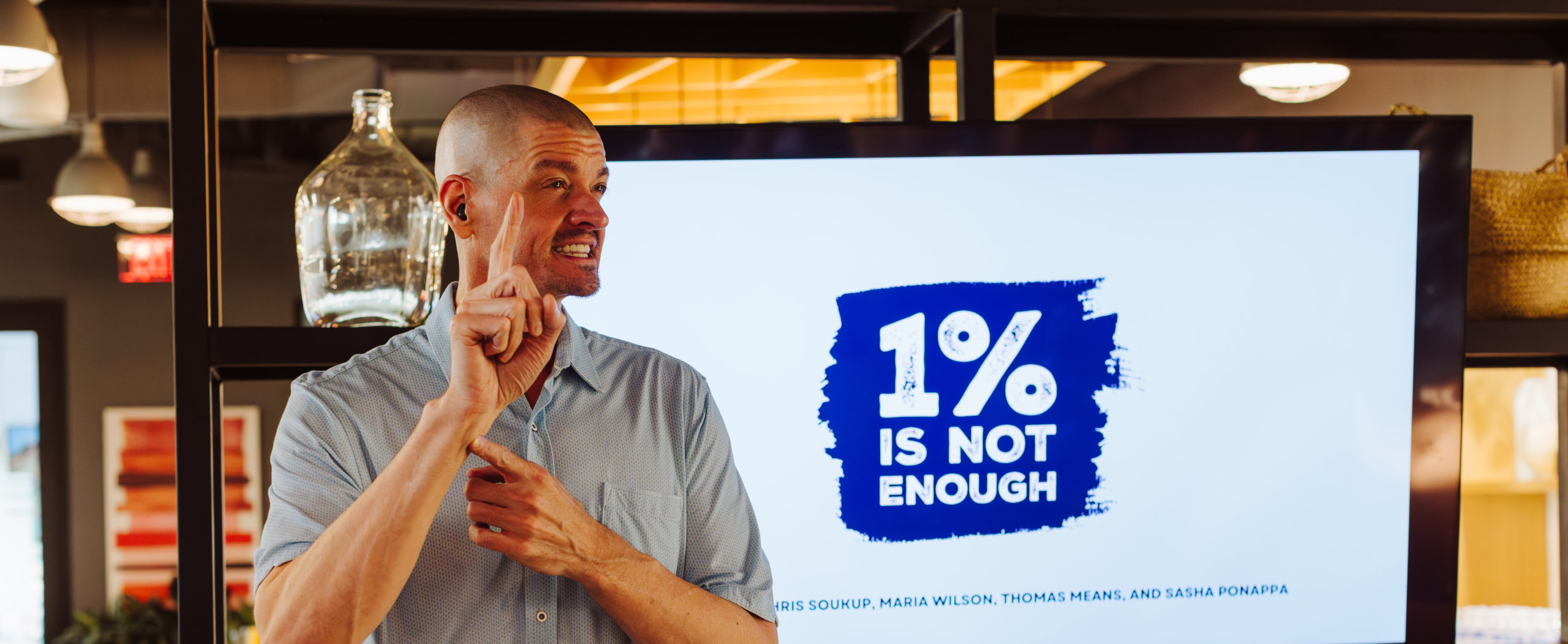 A man with a shaved head signs in front of a large screen that displays the message “1% IS NOT ENOUGH” in bold blue text, inside a modern event space.