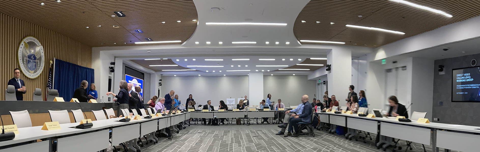 A wide photo shows a large conference room with U-shaped tables, nameplates, and people seated and mingling, with a government seal on the wall and presentation screens visible in the background.