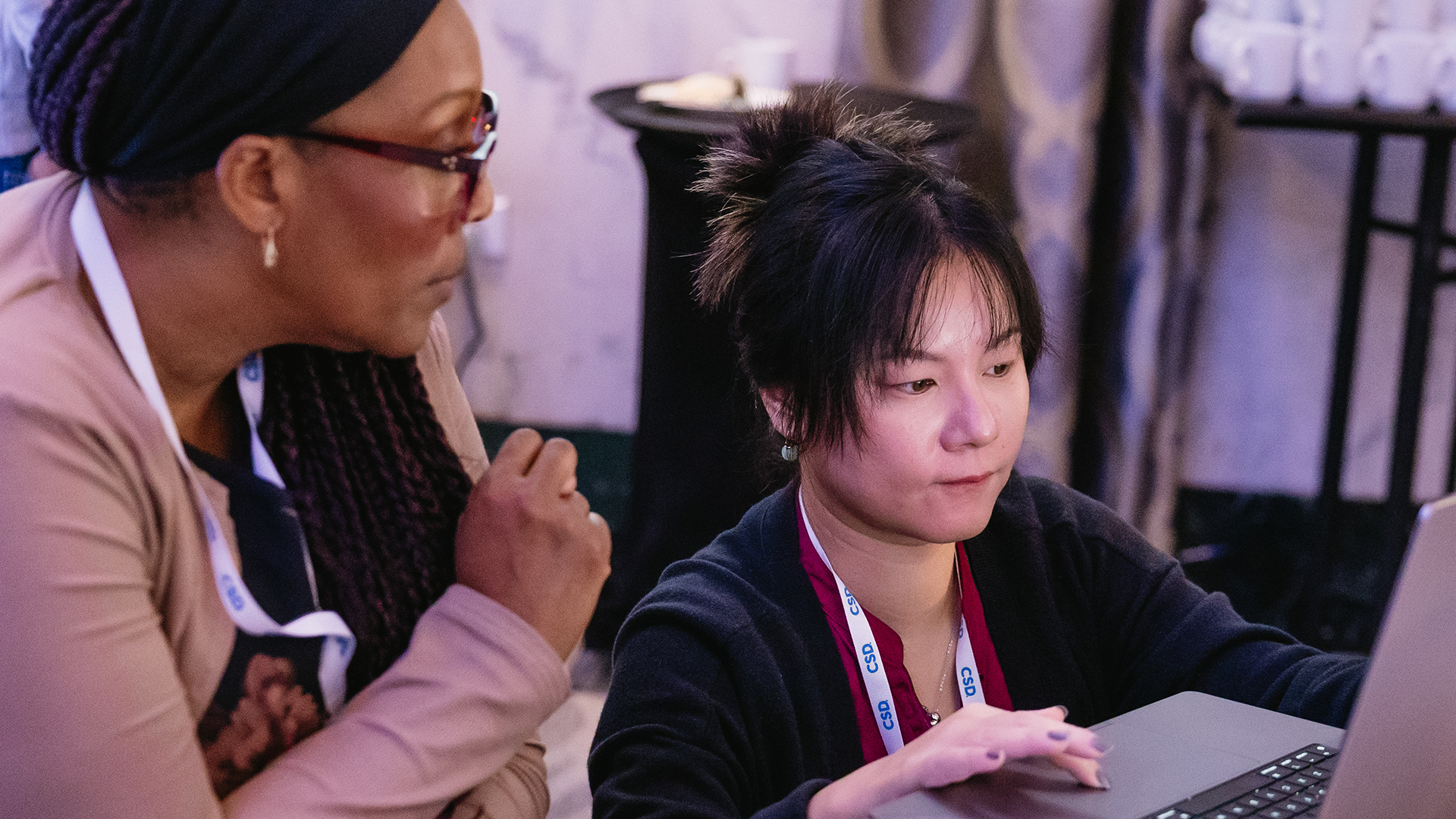 A black woman with braided hair and glasses leans in, watching closely as an Asian woman works on a laptop. Both wear lanyards, and the setting is a workshop space with tables and equipment in the background.