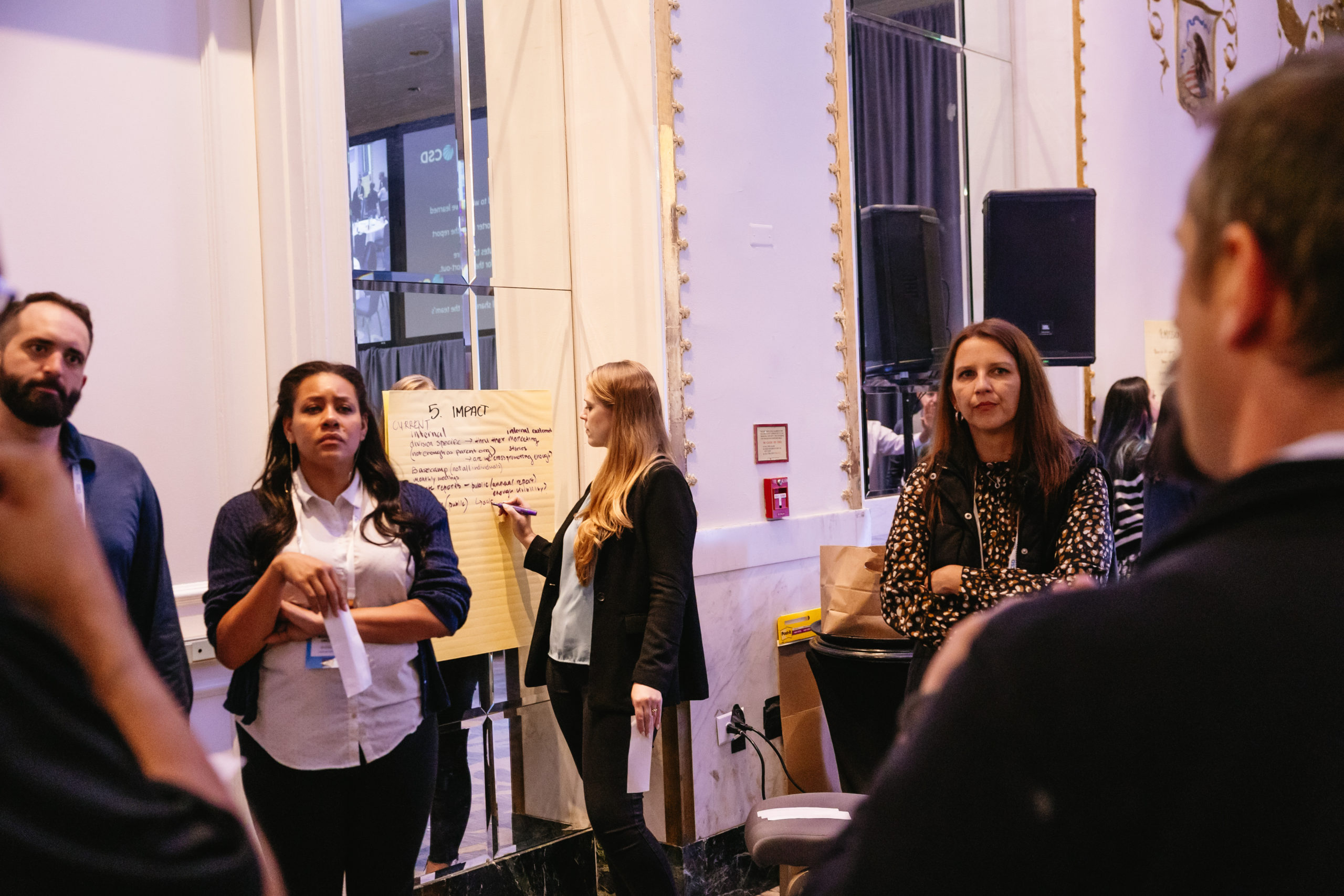A small group of adults stands in a workshop setting. One woman in the center signs, holding a piece of paper, as others watch. Behind her, another woman writes notes on a large flip chart labeled “Impact.” The room has tall mirrors, speakers, and soft lighting, giving it a formal event atmosphere.